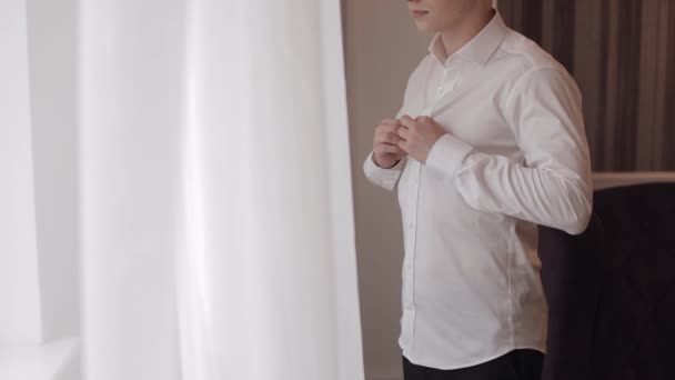 Stijlvolle man in jas knoopt knopen, knoopt witte shirt voor te bereiden om uit te gaan in de buurt van venster — Stockvideo