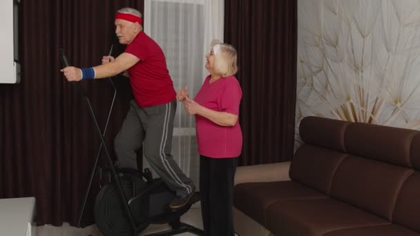 Hombre abuelo maduro activo en ropa deportiva usando orbitrek, la abuela lo anima en casa — Vídeos de Stock