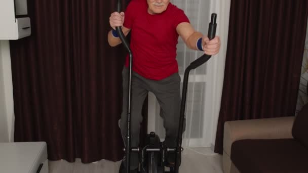 Hombre mayor en ropa deportiva usando orbitrek en la sala de estar haciendo ejercicios de entrenamiento deportivo de cardio en casa — Vídeo de stock