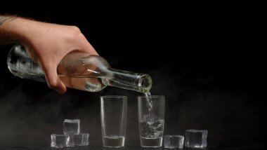 Barmen, siyah arka planda buz küpleri olan iki shot bardağına votka dolduruyor.