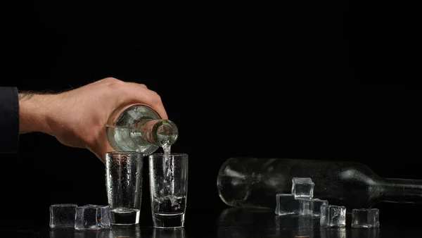 Barman derramando vodka de garrafa em dois copos de tiros com cubos de gelo no fundo preto — Fotografia de Stock