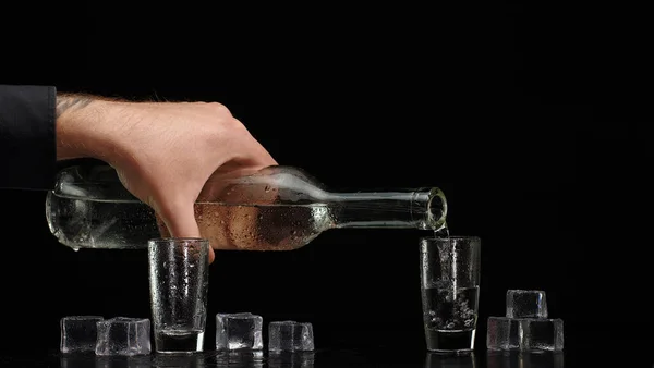 Бармен наливает водку из бутылки в два бокала со льдом кубиками на черном фоне — стоковое фото