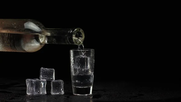 Бармен наливает водку кубиками льда из бутылки в рюмку на черном фоне — стоковое фото