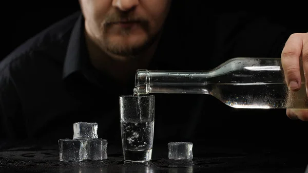 Cantinero vertiendo vodka congelado de botella en vaso de chupito con cubitos de hielo sobre fondo negro — Foto de Stock
