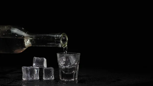 Barman Hand gießt Wodka, Tequila oder Sake aus der Flasche in Glas auf schwarzem Hintergrund mit Eiswürfeln — Stockfoto