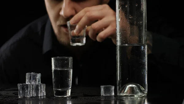 Hombre alcohólico vertiendo vodka congelado de botella en vaso de chupito con cubitos de hielo sobre fondo negro — Foto de Stock