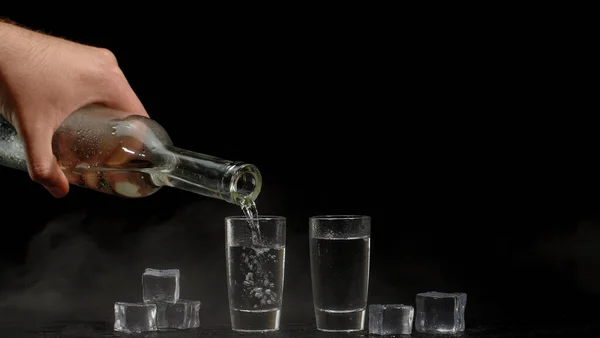 Рука наливает водку, текилу или сакэ из бутылки в рюмки на черном фоне с кубиками льда — стоковое фото