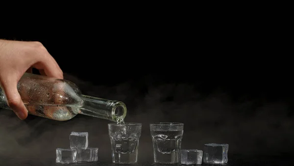 Бармен наливает две рюмки водки с кубиками льда из бутылки в очки на черном фоне — стоковое фото