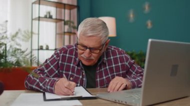 Dizüstü bilgisayarda çalışan kıdemli bir adam, kâğıt üzerinde notlar alıyor, kötü sonuçlara üzülüyor.