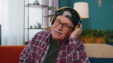 Kablosuz kulaklıklı mutlu adam dans ediyor, evdeki oturma odasında rahat bir kanepede şarkı söylüyor.