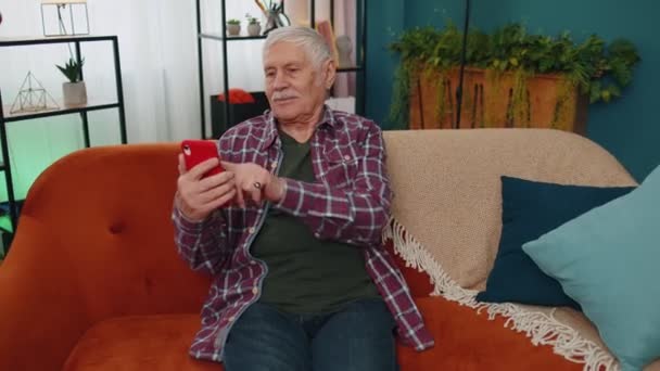 Abuelo de edad avanzada trabaja en el teléfono móvil, envía mensajes, hace compras en línea en el sofá del hogar — Vídeos de Stock
