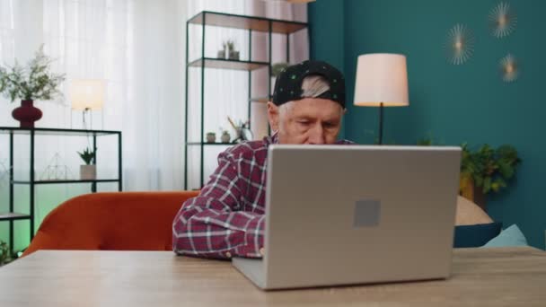 Abuelo mayor usando computadora portátil, escribiendo en la habitación de casa, mirando a la cámara, sonriendo — Vídeo de stock