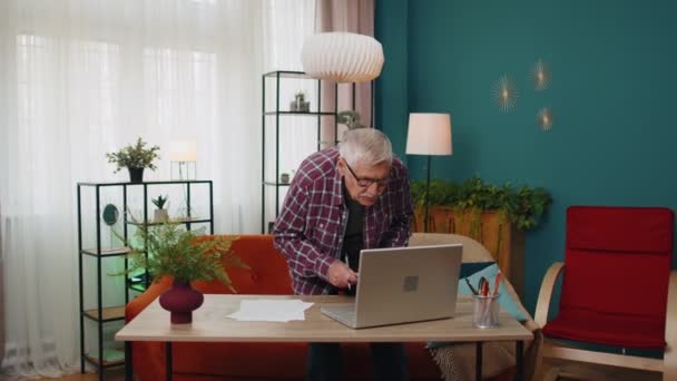 Старший дедушка, работающий на ноутбуке, делает заметки на листе бумаги, рассчитывая счета — стоковое видео