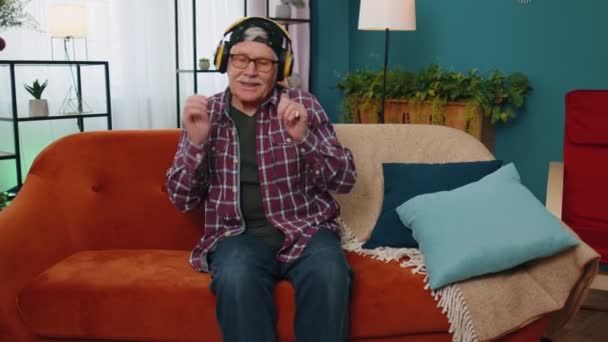 Homem idoso muito feliz em fones de ouvido sem fio dançando, cantando no sofá acolhedor na sala de estar em casa — Vídeo de Stock