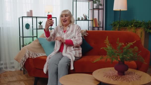 Счастливая старшая бабушка со смартфоном смотрит мобильное видео и делает селфи дома — стоковое видео