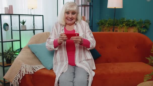 Preocupado divertida abuela mujer con entusiasmo jugando videojuegos de carreras en el teléfono móvil en casa — Vídeos de Stock