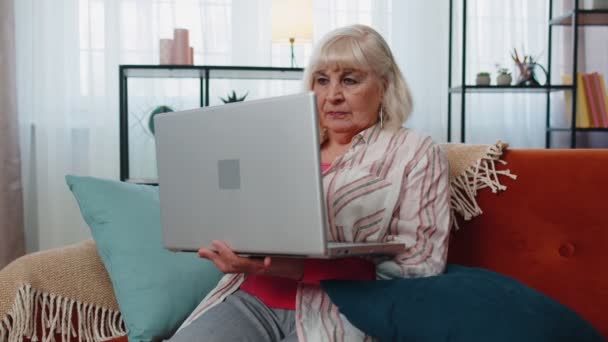 Starsza babcia korzystająca z laptopa, pisząca w domu, patrząca w kamerę, uśmiechnięta — Wideo stockowe