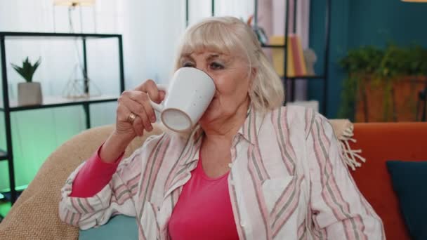 Senior stylowy stary babcia kobieta siedzi na kanapie relaks w domu picia cieszyć się herbaty lub kawy — Wideo stockowe