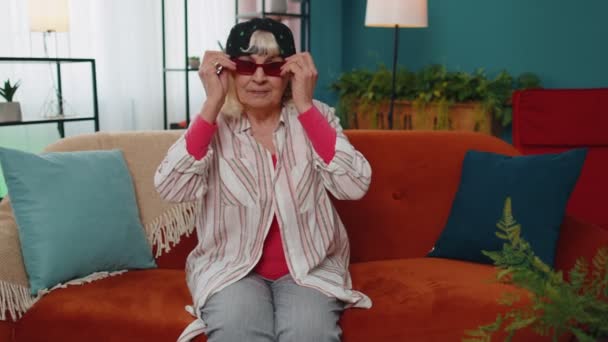 Κομψή γιαγιά με καπέλο μαριχουάνας, φοράει κόκκινα γυαλιά ηλίου, κοιτάζει την κάμερα με γοητευτικό χαμόγελο — Αρχείο Βίντεο