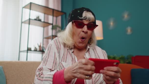Preocupado divertida abuela mujer con entusiasmo jugando videojuegos de carreras en el teléfono móvil en casa — Vídeo de stock