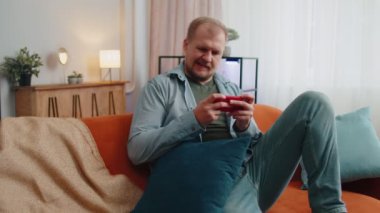 İnternette video oyunu oynayan endişeli, komik yetişkin adam evde cep telefonuyla kazanmayı kutluyor
