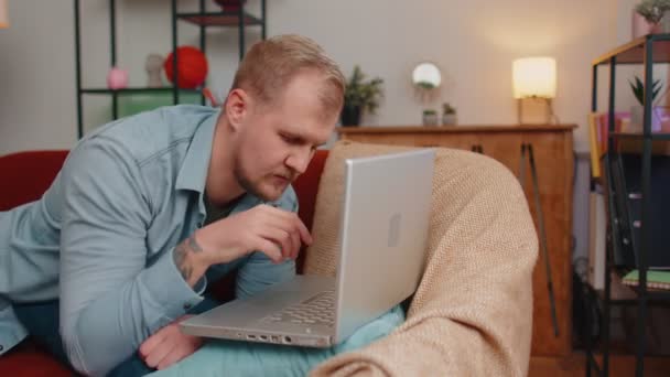 Yetişkin bir adam dizüstü bilgisayar kullanıyor. Evdeki koltukta yatıyor, çalışıyor, online alışveriş yapıyor, film izliyor. — Stok video