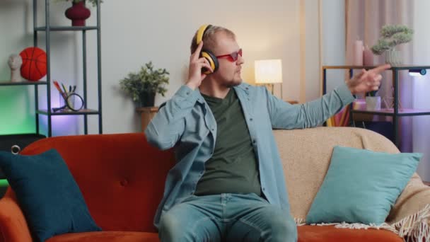 Överlycklig ung man i trådlösa hörlurar dansar, sjunger på mysig soffa i vardagsrummet hemma — Stockvideo