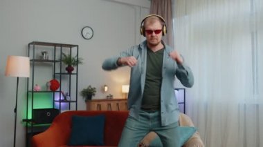 Kablosuz kulaklıklı neşeli genç adam dans ediyor, evdeki oturma odasında rahat koltukta şarkı söylüyor.