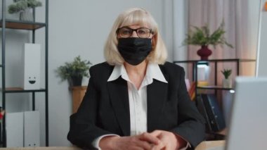 Üst düzey çalışan yüz maskesi takıyor. Ofisten tek başına çalışıyor. Masada oturup kameraya bakıyor.