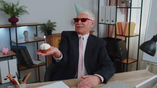 Старший бізнесмен святкує самотній день народження в офісі, дме свічку на маленькому торті, роблячи бажання — стокове відео