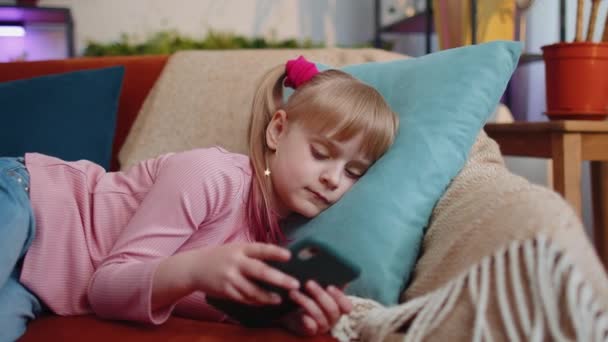 Dziecko maluch trzymać smartfon oglądając śmieszne kreskówki, czatując z przyjaciółmi na kanapie sam w domu — Wideo stockowe