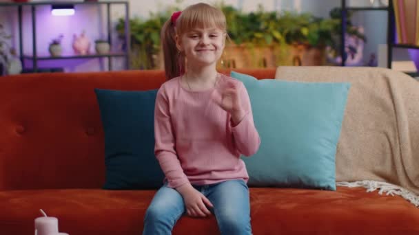 Child girl sitting on sofa at home looking at camera smiling waving hands gesturing hello or goodbye — Stockvideo