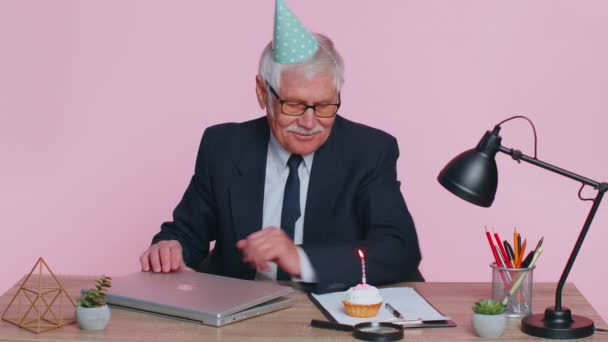 Starszy biznesmen świętuje samotne urodziny w biurze, dmuchając świeczkę w małe ciasteczko wypowiadając życzenie — Wideo stockowe