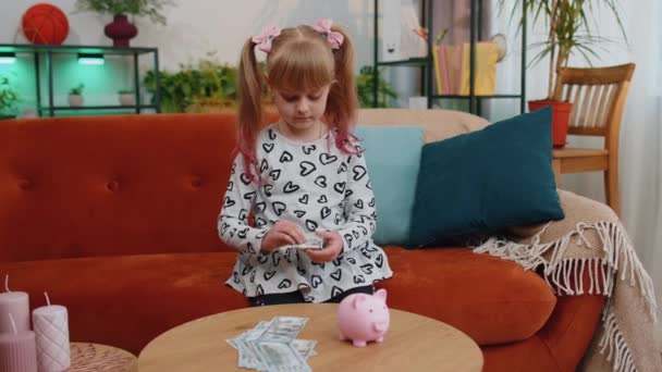 Little toddler kid girl putting money dollar banknotes for future gift purchase at piggybank at home — Αρχείο Βίντεο