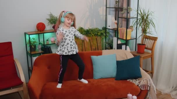 Glückliche Kindermädchen mit Kopfhörern tanzen und springen, während sie allein zu Hause Musik hören — Stockvideo