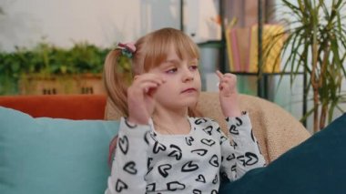 Little toddler girl relaxing on sofa, wearing wireless earphones, listening to music, favorite song