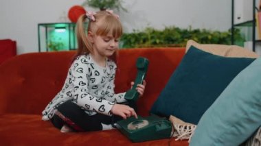 Smiling young children girl making conversation call on retro phone with friends on couch at home