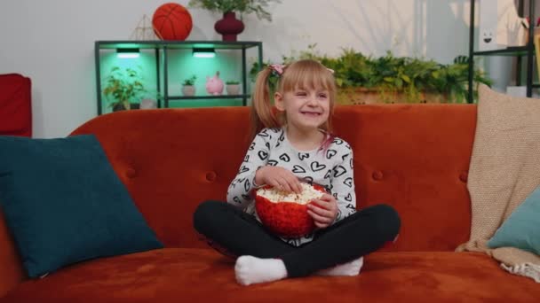 Funny children girl watching comedy video film on tv, eating popcorn on comfortable sofa at home — Vídeos de Stock