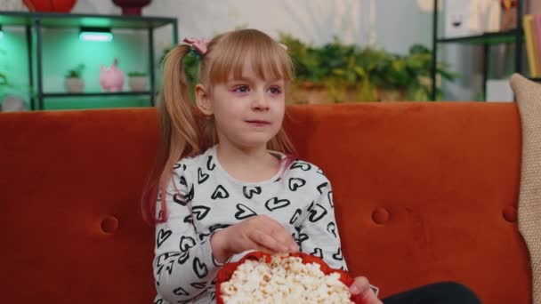 Смішна маленька дівчинка дивиться комедійний відеофільм на телебаченні, їсть попкорн на зручному дивані вдома — стокове відео