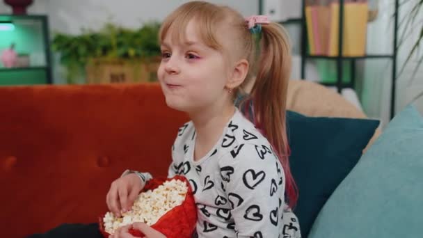 Funny children girl watching comedy video film on tv, eating popcorn on comfortable sofa at home — Stok video