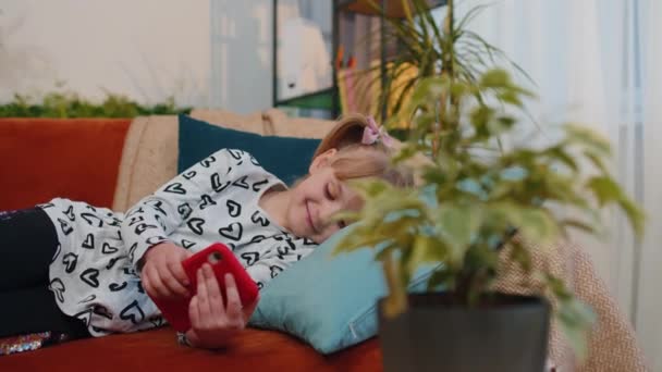 Niño niño sostiene teléfono inteligente viendo dibujos animados divertidos, charlando con amigos en el sofá solo en casa — Vídeos de Stock