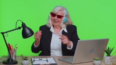 Senior business woman listening music on headphones, dancing disco fooling around, celebrating win