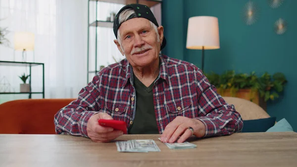 Sorrindo homem idoso segurando dinheiro em dinheiro e smartphone, satisfeito com a renda, planejamento do orçamento familiar — Fotografia de Stock