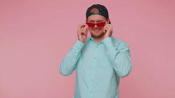 Seducente allegro uomo elegante in camicia blu con occhiali da sole, sorriso affascinante sulla parete rosa — Foto Stock