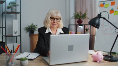 Gülümseyen olgun iş kadını ofis masasına oturur, dizüstü bilgisayarı açar ve çalışmaya başlar.