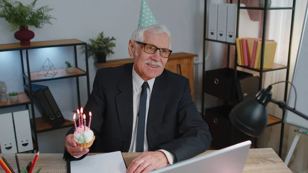Senior old business man celebrating birthday in office holding small cake with candles making a wish