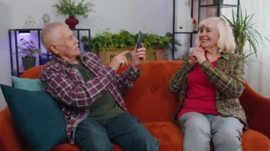 Sosyal medya için akıllı telefondan fotoğraf çeken yaşlı, yaşlı bir çift, büyükanne ve büyükbaba kadın. Olgun büyükbaba blogcuları cep telefonu kullanıyor. Evdeki koltukta poz veriyor ve gülümsüyor.