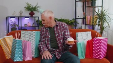 Yaşlı ve mutlu yaşlı bir adam neşeli bir doğum günü şapkası takıyor ve kekin üzerine yanan mum üflediği için mutlu bir dilek tutuyor. Olgun büyükbaba partisini evde hediyelerle kutluyor.