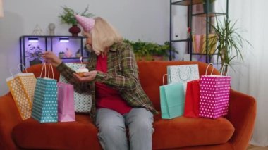 Yaşlı ve mutlu yaşlı bir kadın neşeli bir doğum günü şapkası takıyor ve kek tutarken pastaya mum üflediği için sevinçten havalara uçuyor. Olgun büyükanne evde hediyelerle koltukta parti veriyor.