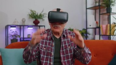 Kıdemli yaşlı adam, simülasyon oyunu oynamak için kulaklık uygulaması kullanıyor, simülasyon gözlüğü cihazı teknolojisi. Sanal gerçeklik 3D 360 videosu izliyoruz. Evde kanepede VR gözlüklü yetişkin büyükbaba.